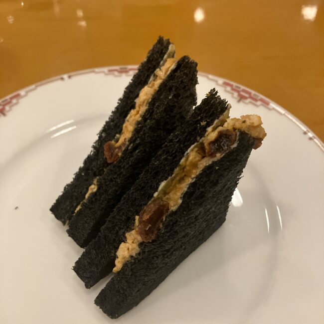 帝国ホテル アフタヌーンティーの竹炭サンドイッチの断面。黒いパンの間に、カレー風味のチキンとレーズンのフィリングが挟まっている。