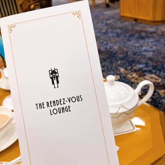 帝国ホテル 東京「ランデブーラウンジ」のメニュー表紙。白地に金色の枠線とアールデコ調のロゴ、"THE RENDEZ-VOUS LOUNGE"の文字が記されている。奥には白いティーポットとラウンジの特徴的な青いカーペットが見える。