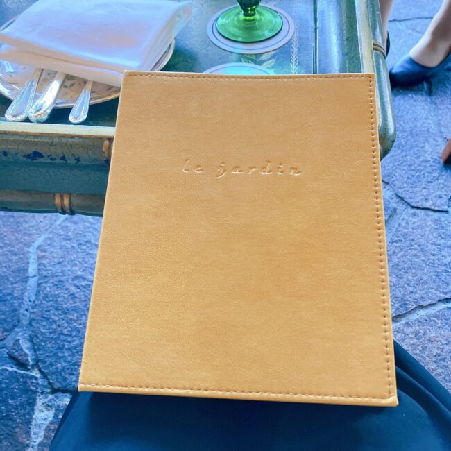 ホテル椿山荘東京「ル・ジャルダン」のアフタヌーンティー。キャメル色のレザー素材のメニューブックが、大理石調のテーブルの上に置かれている。中央には「le jardin」のロゴがエンボス加工されている。背景にはカトラリーや白いナプキンが見え、優雅なラウンジの雰囲気が伝わる。