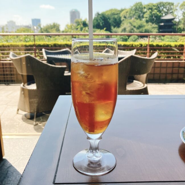 ホテル椿山荘東京「ル・ジャルダン」のアフタヌーンティー。テラス席で提供された、背の高いステムグラス入りのアイスティー。グラスの中の氷は透明ではなく、紅茶と同じ深い琥珀色をしており、氷自体も紅茶で作られているのが視覚的に確認できる。背景には豊かな庭園と都会のスカイラインが見える。