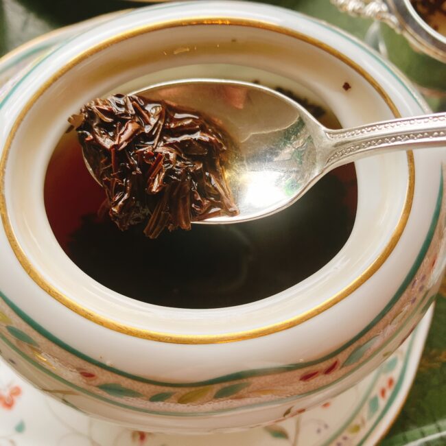 ノリタケ「花更紗」のティーポットに直接入った茶葉を、スプーンですくっているクローズアップ。ティーバッグではなく茶葉から抽出する本格英国スタイルを示す。