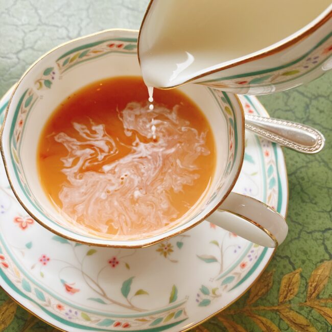 ノリタケ「花更紗」のティーカップに入った紅茶に、クリーマーからミルクが注がれている。ミルクが紅茶の中でマーブル状に広がっている瞬間。
