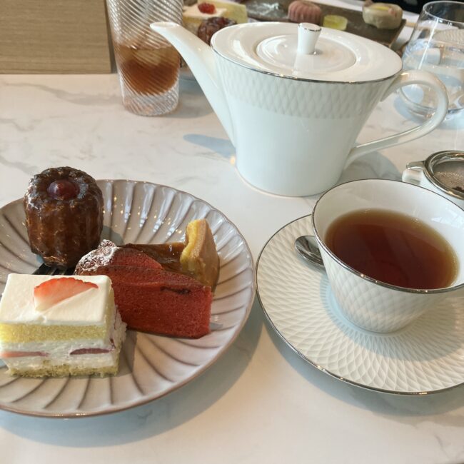 白いティーポットと、紅茶が注がれたティーカップ。手前には、カヌレ、ショートケーキ、赤いパウンドケーキなどが盛られた皿。