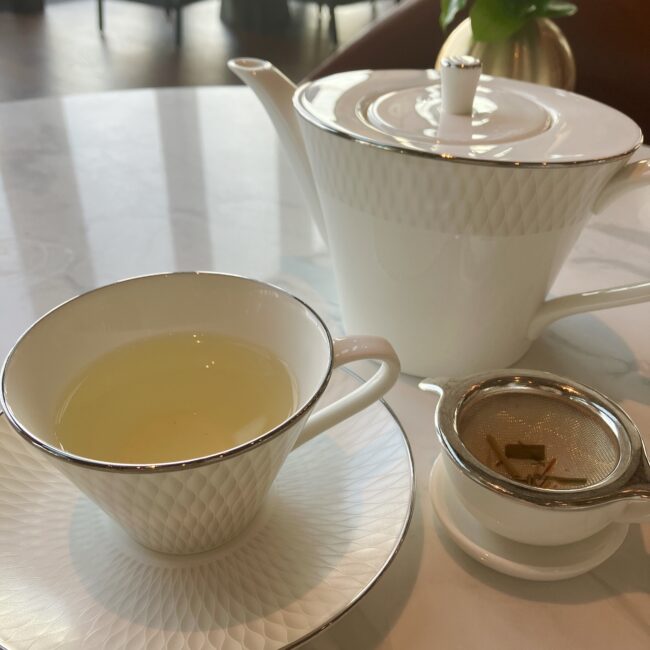 白いティーポットから、白いティーカップへ注がれる、淡い黄色のハーブティー。茶漉しにはレモングラスなどのハーブが見える。