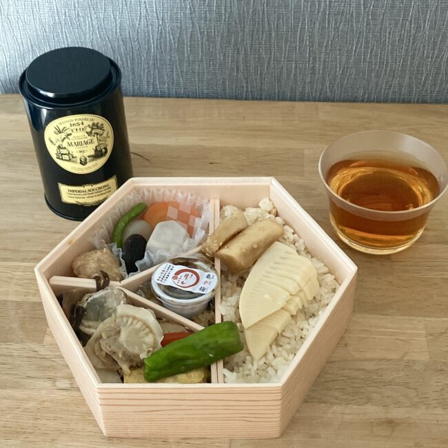 マリアージュフレールの紅茶「インペリアル スーション (IMPERIAL SOUCHONG)」の黒缶と、ガラスカップに注がれた琥珀色の紅茶。手前には、桝本の六角形の木製お弁当（竹の子ご飯や煮物など）が並んだ、和食とのペアリング写真。