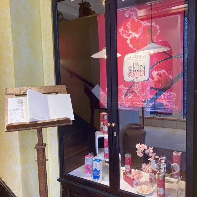 マリアージュフレール銀座松屋通り店の入り口。黒いショーケースには、鮮やかなピンクの「サクラ, サクラ! 2026」の特大ポスターと、桜のパッケージの茶筒、ティーポットが美しくディスプレイされている。左手にはメニュー・スタンドがある。