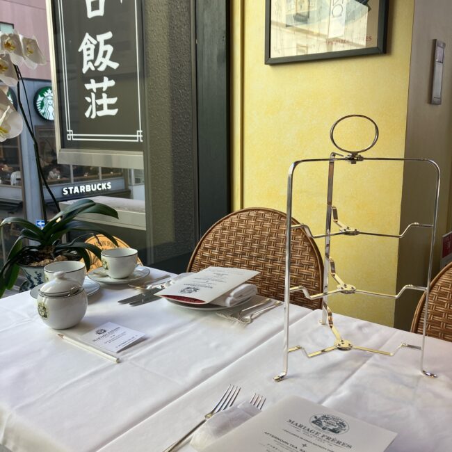 マリアージュフレール銀座松屋通り店のサロン・ド・テ窓際席。白いテーブルクロスの上に、ティーカップ、シュガーポット、カトラリー、メニュー表、そしてシルバーの3段ティースタンド(空の状態)が美しくセットされている。窓の外には向かいのスターバックスが見える。