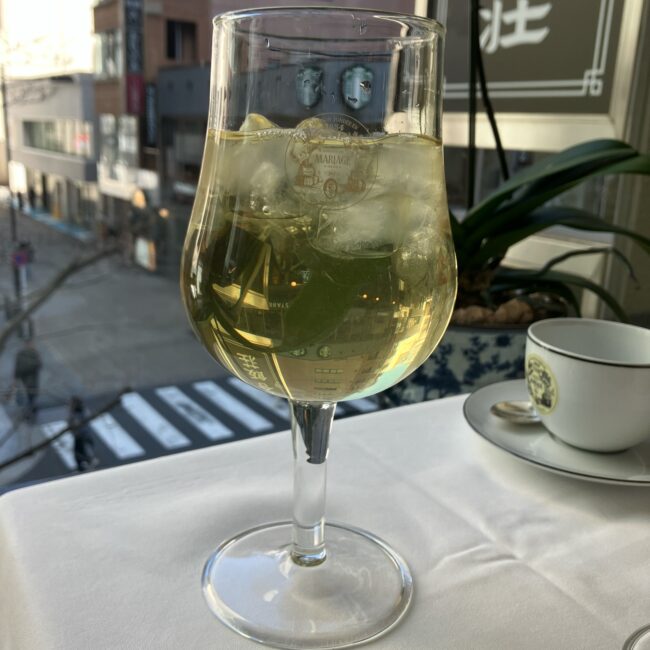 銀座の街並みとスターバックスのロゴが見える窓を背景に、マリアージュフレールのロゴ入りグラスに注がれたグリーンティーのアイスティー。氷がたっぷり入った爽やかな様子