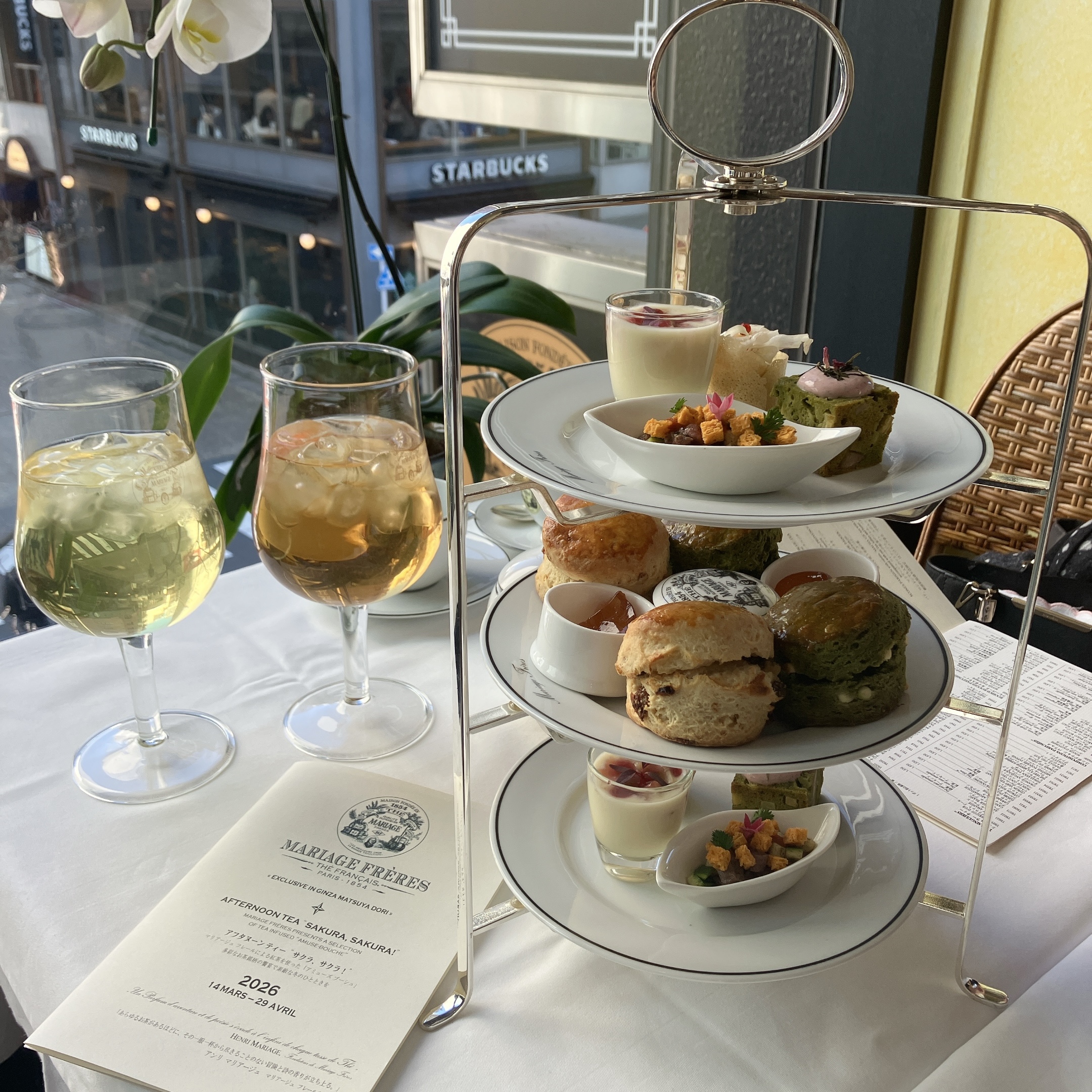 マリアージュフレール銀座松屋通り店のアフタヌーンティー「サクラ、サクラ! 2026」の豪華な3段シルバースタンド。中段には抹茶スコーン、上段と下段には桜色のスイーツが並ぶ。手前には2つの大きなアイスティーグラスと、サクラアフタヌーンティー2026のメニューが置かれている。窓の外にはスターバックスが見える