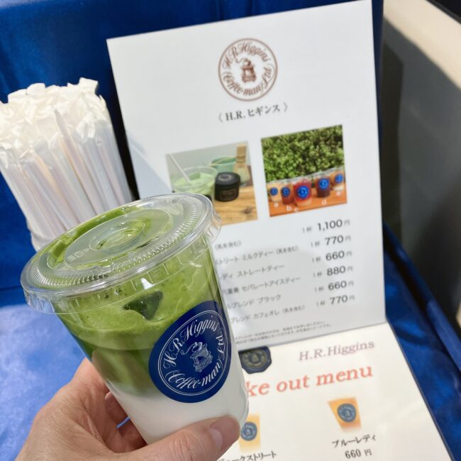 H.R.ヒギンズのテイクアウト用抹茶ラテ。背景にはメニュー表。