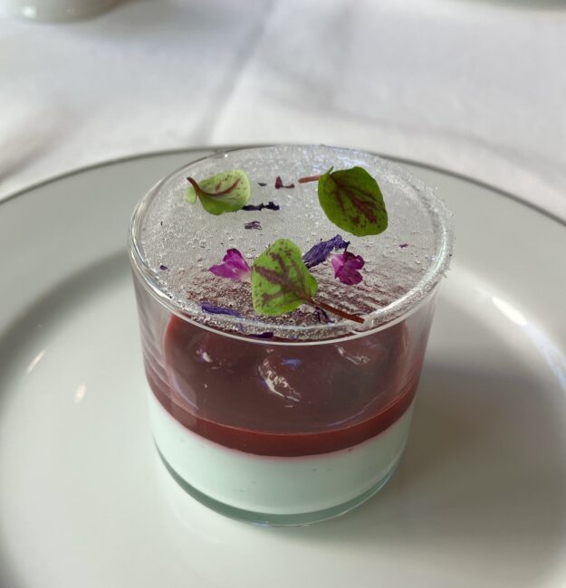 マリアージュフレールのアフタヌーンティーのスイーツ。「青茶桜ブルーティー はなやぐパンナコッタ」。グラスに入ったパンナコッタの上に、薄い氷のような砂糖細工、葉、紫の花が乗っている。