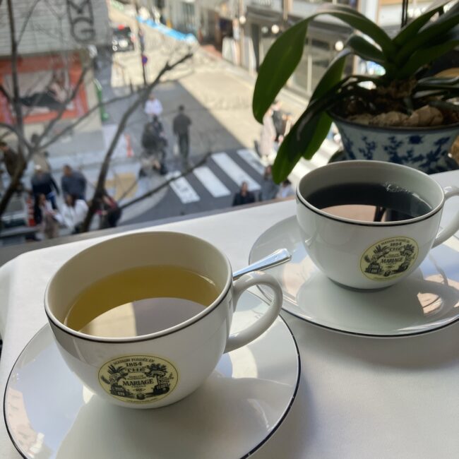 マリアージュフレール銀座松屋通り店のサロン・ド・テ窓際席。白いテーブルクロスの上に、マリアージュフレールのロゴ入りティーカップ2客が並び、それぞれ淡い黄緑色の緑茶と、濃い褐色の紅茶が注がれている。窓の外には向かいのスターバックスが見える。