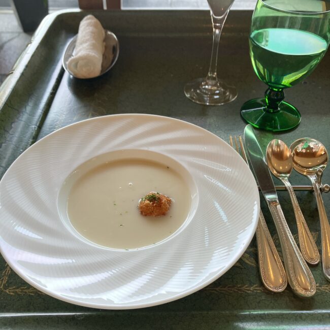 ホテル椿山荘東京「ル・ジャルダン」の桜＆ストロベリーアフタヌーンティーで提供されるアミューズ。白いスープ皿に入ったカブのブラマンジェに、小さな鯛ブランダードのコロッケとパルマンティエが添えられている。緑色のグラスやカトラリーと共にトレーに乗せられている様子。