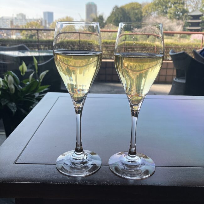 ホテル椿山荘東京「ル・ジャルダン」の桜＆ストロベリーアフタヌーンティーのウエルカムドリンク。テラス席のテーブルに、薄黄色のドリンクが注がれた2つのシャンパングラスが置かれている。背景には、緑豊かな庭園と三重塔、ホテル棟が見える。