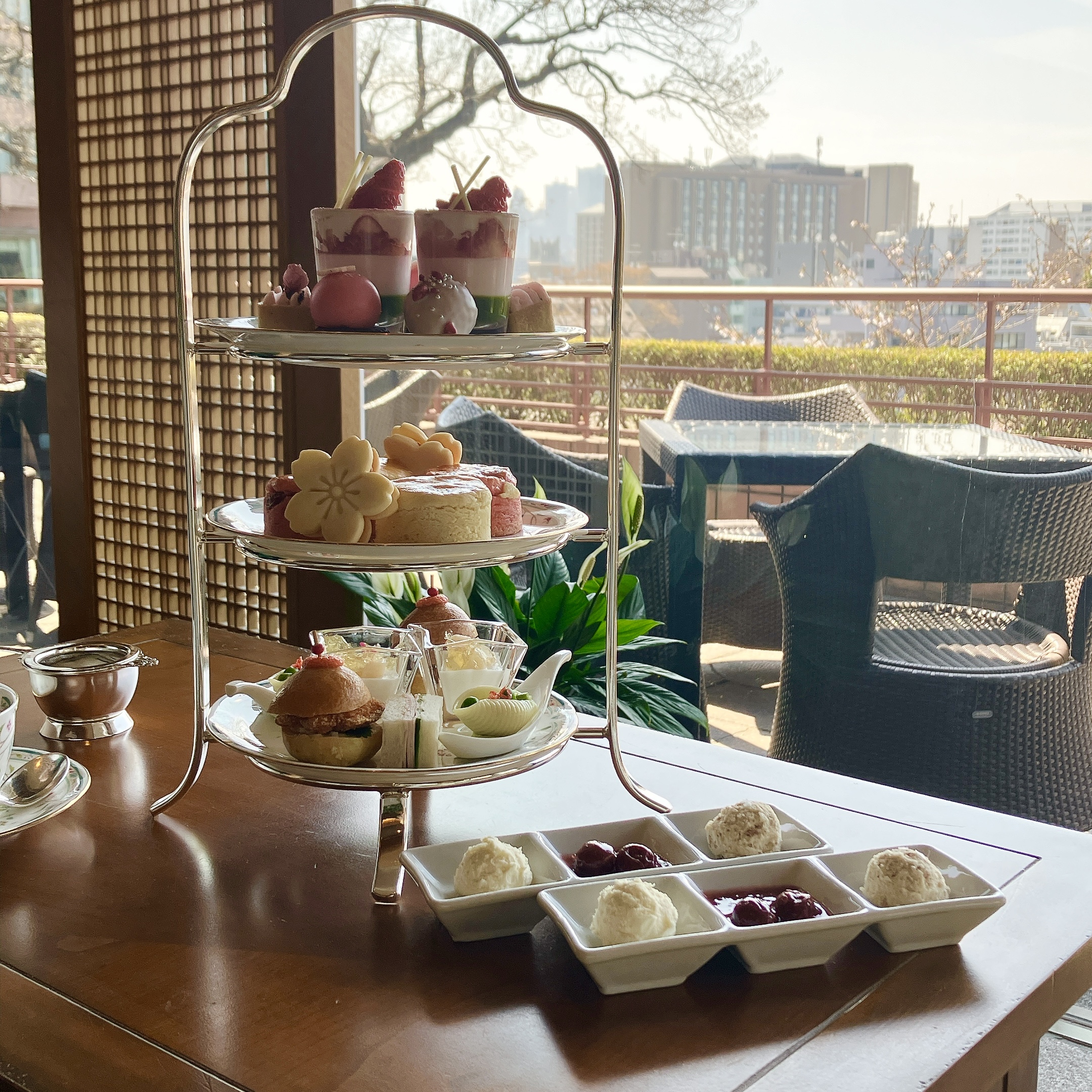 ホテル椿山荘東京「ル・ジャルダン」の2026年桜＆ストロベリーアフタヌーンティー。窓際のテーブルに置かれた3段スタンドの全体像。上段に苺と桜のスイーツ、中段にスコーンと桜クッキー、下段にアミューズを含むセイボリーが並び、手前にコンディメントが添えられている。背景には緑豊かな庭園が広がる。