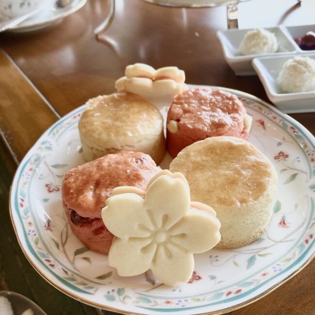 アフタヌーンティースタンドの中段（スコーン）。ウェッジウッド風の柄の皿に、プレーンといちごの2種類のスコーンが各2つ、その手前にピンク色の桜の形のクッキーが1つ置かれている。