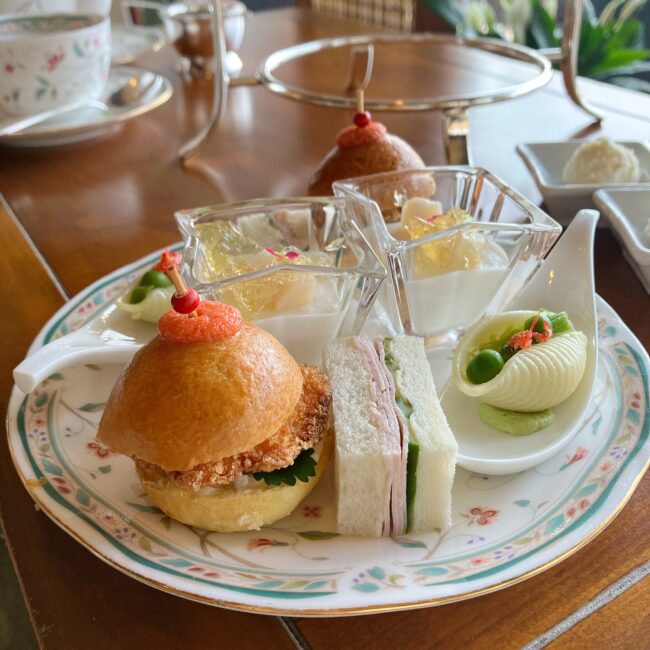 アフタヌーンティースタンドの下段（セイボリー）。同じ柄の皿に、鯛ブランダードのコロッケ付きフィッシュバーガー、自家製ハムときゅうりのティーサンド、カブのブラマンジェ、コンキリエのアーリオ・オーリオが並んでいる。