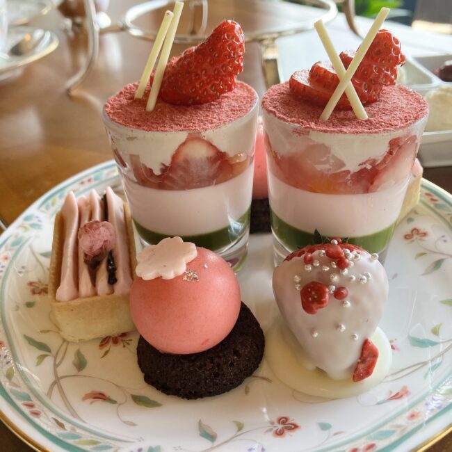 アフタヌーンティースタンドの上段（スイーツ）。銀のスタンドに乗ったガラス皿に、苺と桜のヴェリーヌが2つ、ホワイトチョコレートがかかった苺、ピンク色の球形ムース、ピンク色のモンブランタルトが並んでいる。