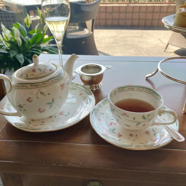 ノリタケ「花更紗」シリーズのティーポットと、紅茶が入ったティーカップ＆ソーサー。奥にはティーストレーナー（茶こし）が添えられている。窓際のテーブルでの優雅なセット。