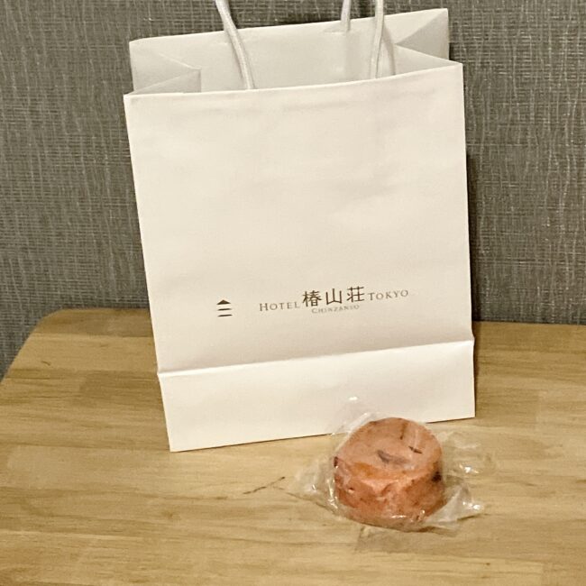 自宅のテーブルに置かれた、ホテル椿山荘東京のロゴ入り紙袋。手前に個包装されたピンク色のスコーン（イチゴ）が1個置かれている。