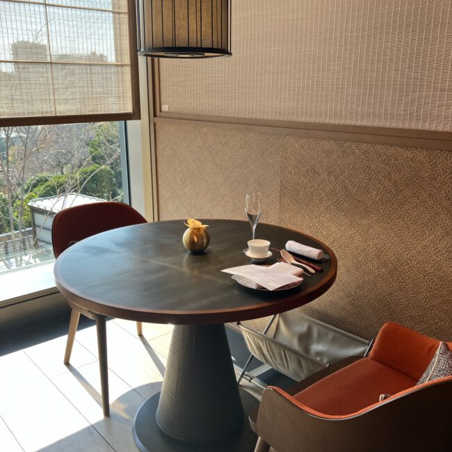 ジャヌ東京「虎景軒」の窓際のテーブル席の全体像。日差しが差し込む窓辺に、丸いテーブルとオレンジ色の椅子が2脚配置され、テーブルセットが用意されている。