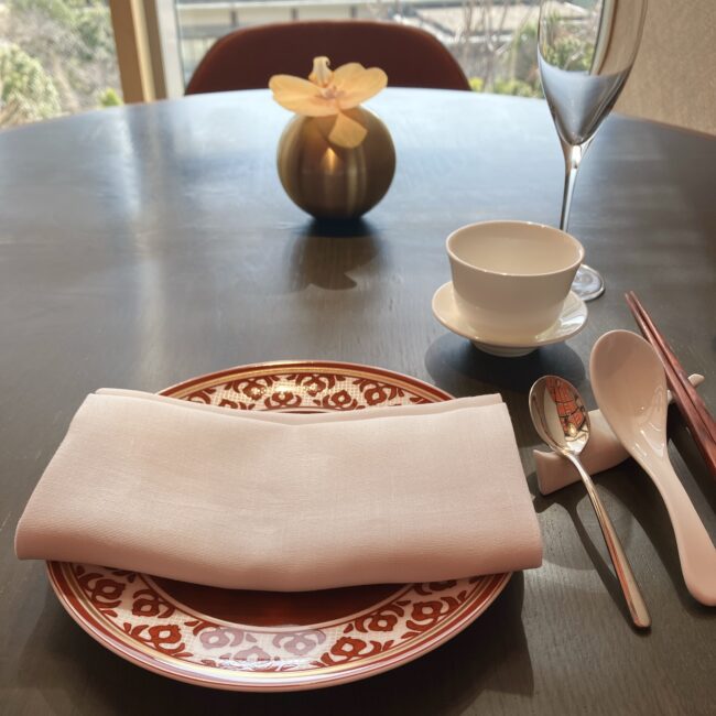 ジャヌ東京「虎景軒」の窓際のテーブル席。赤い縁取りの中華皿、ナプキン、お箸、レンゲ、スープスプーン、ティーカップ、蘭の花が活けられた金色の花瓶、シャンパングラスがセットされている。