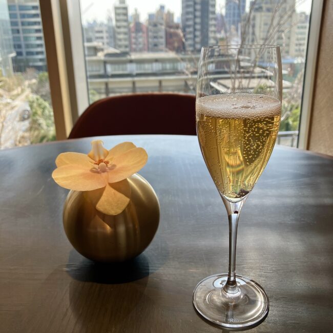 ジャヌ東京「虎景軒」の窓側の席。金色の花瓶に活けられた蘭の花の隣に、ウェルカムドリンクのスパークリングワインが注がれたシャンパングラスが置かれている。窓の外には東京の街並みが広がる。