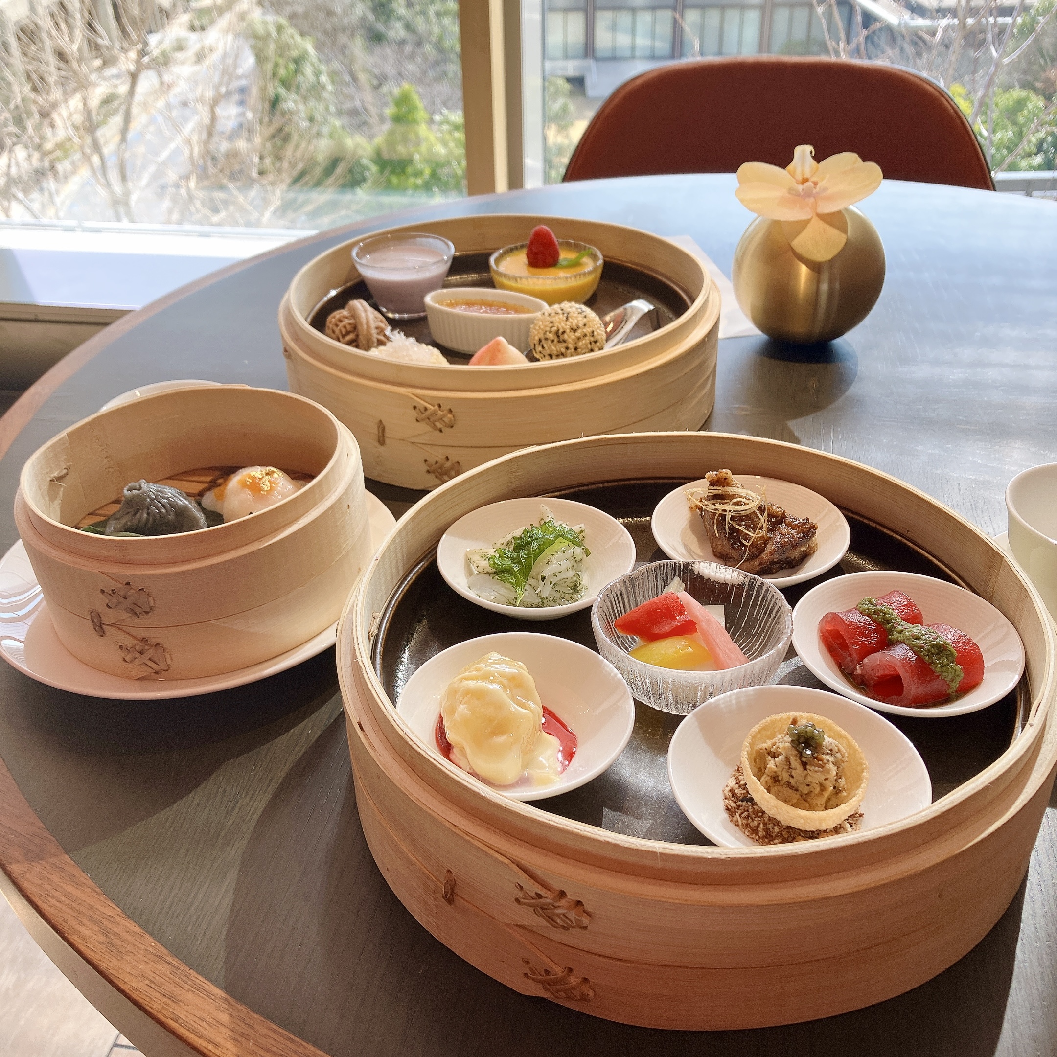 麻布台ヒルズ「ジャヌ東京」の中国料理「虎景軒（フージン）」で提供される、1日10食限定の中華アフタヌーンティーの全体写真。自然光が差し込む窓際のテーブルに、せいろに盛られた燕の巣、和牛、北京ダックなどの豪華な点心やスイーツが並んでいる様子。