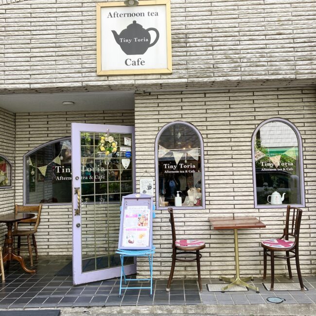 都営浅草線・日比谷線「人形町駅」A2出口から徒歩2分。「甘酒横丁」の近くにある「タイニートリアティールーム」の可愛らしい英国風の外観。白いレンガ調の壁に、特徴的な薄紫色のドア、アーチ型の窓、店頭のテラス席が見える。