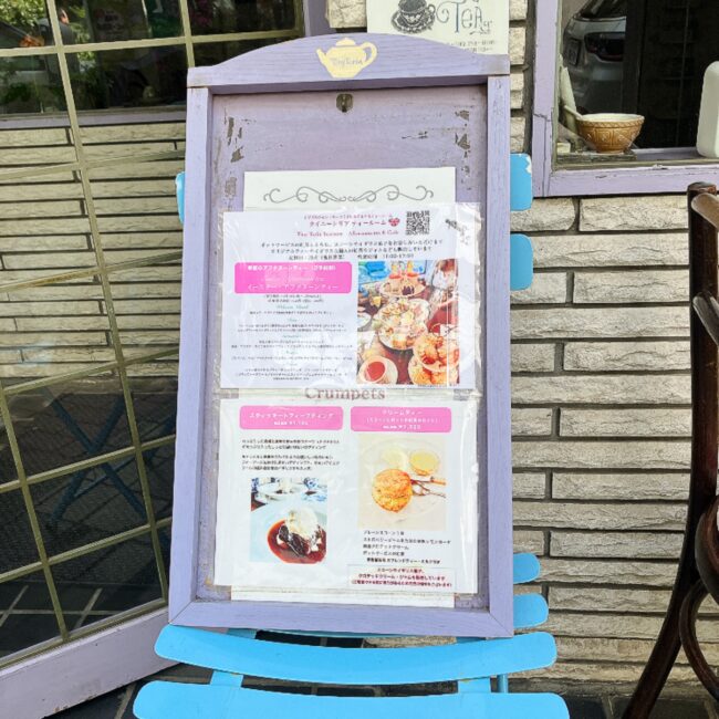 タイニートリアティールームの店頭に置かれた紫色のイーゼル看板。期間限定の「イースターアフタヌーンティー」のメニュー内容や、食べきれないスコーンの持ち帰り、その他メニュー（クランペット、クリームティー）についての案内が記載されている。