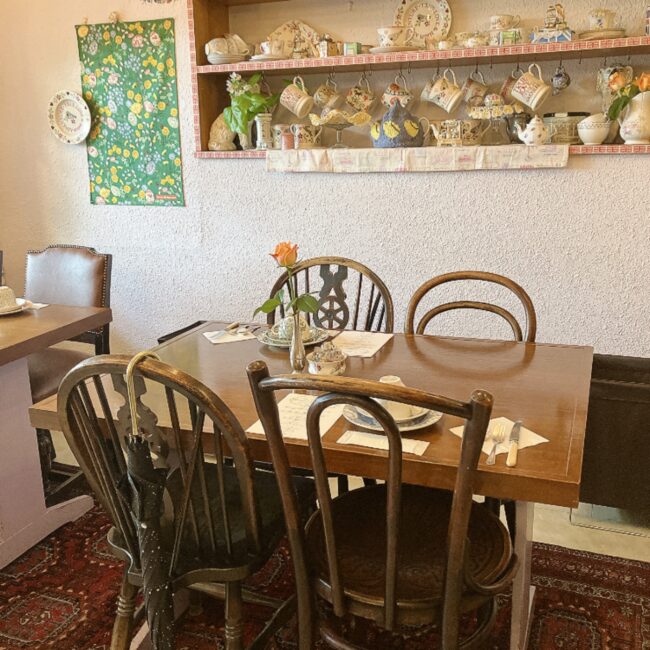 タイニートリアティールームの店内。イギリス製のアンティークな木製テーブルと椅子が並び、壁際の棚には可愛らしいティーカップやティーポットがずらりと飾られている温かみのある空間。