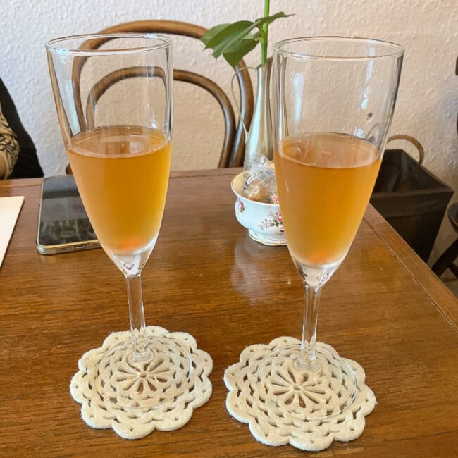 タイニートリアのウェルカムドリンク「春摘みダージリンと大島桜のアイスティー」。細長いシャンパングラスに注がれており、グラスの底には桜の花びらが沈んでいる。白いレース編みのコースターに乗せられ、アンティークな木製テーブルに置かれている。