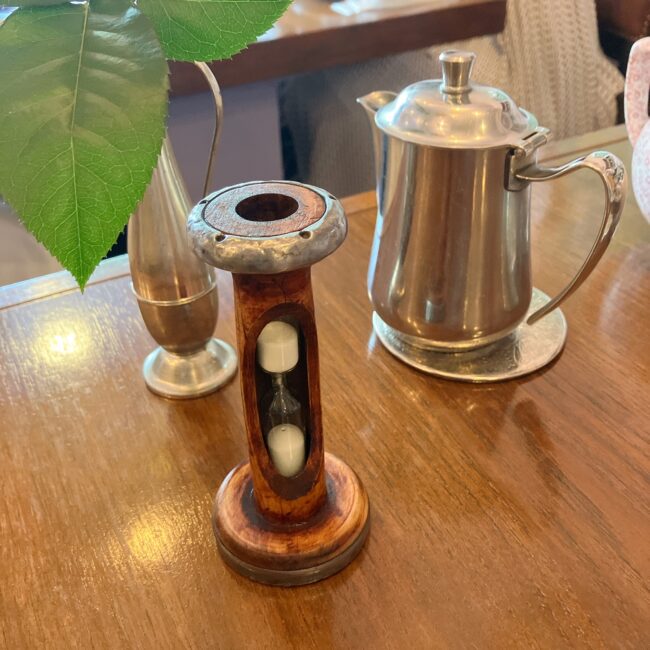 タイニートリアのテーブルに置かれた、紅茶の抽出時間を計るためのアンティーク調の木製砂時計。隣には差し湯用のシルバーポットが並んでおり、本格的なイギリス式ティーサービスを楽しめる。