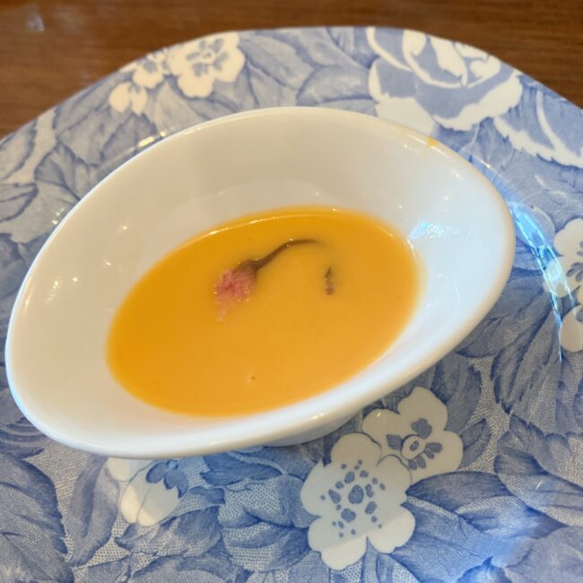 タイニートリアのイースターアフタヌーンティーのセイボリー。「桜と人参の冷製スープ」。鮮やかなオレンジ色のスープの中心に、春らしい桜の花びらが浮かべられている。