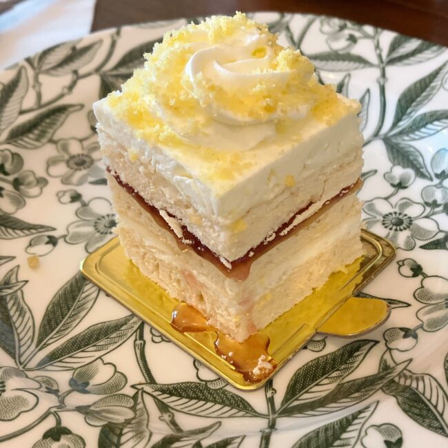 タイニートリアのスイーツ「メレンゲミモザケーキ」。スポンジとジャム、クリームが層になり、トップにはミモザの花に見立てた黄色のスポンジクラムとホイップクリームが華やかに飾られている。緑色のボタニカル柄のプレートによく映えている。