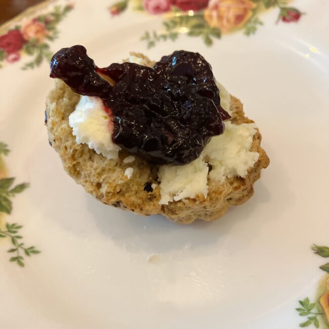 半分に割った温かいイースタービスケットスコーン。その上に、真っ白なクロテッドクリームと、果肉感たっぷりのブルーベリージャムが山盛りに乗せられている。花柄の美しいプレートの上にある様子。