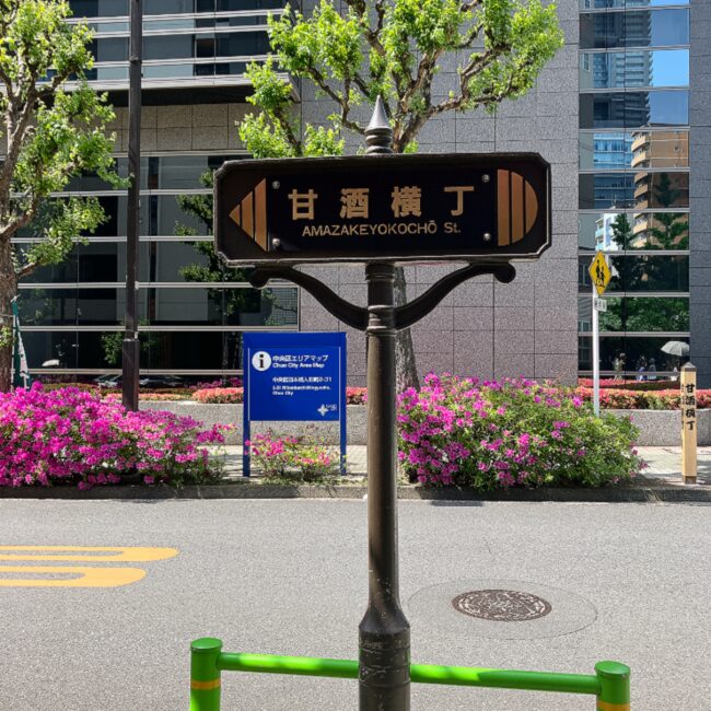 人形町の歴史ある通り「甘酒横丁（Amazake yokocho St.）」の茶色の標識。背景には美しいピンク色のツツジが満開に咲いており、その先にタイニートリアティールームが位置する。