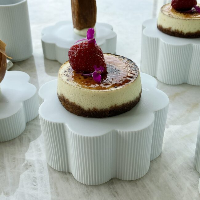 クリーミーチーズケーキ スペキュロスクッキーベース。表面がブリュレされており、上に新鮮なラズベリーと紫の花が飾られている。