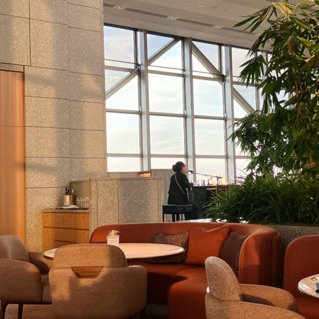夕暮れ時のパークハイアット東京「ピーク ラウンジ」。41階の窓から差し込む夕日を背景に、生演奏をするギタリストの後ろ姿と、落ち着いた雰囲気の店内。
