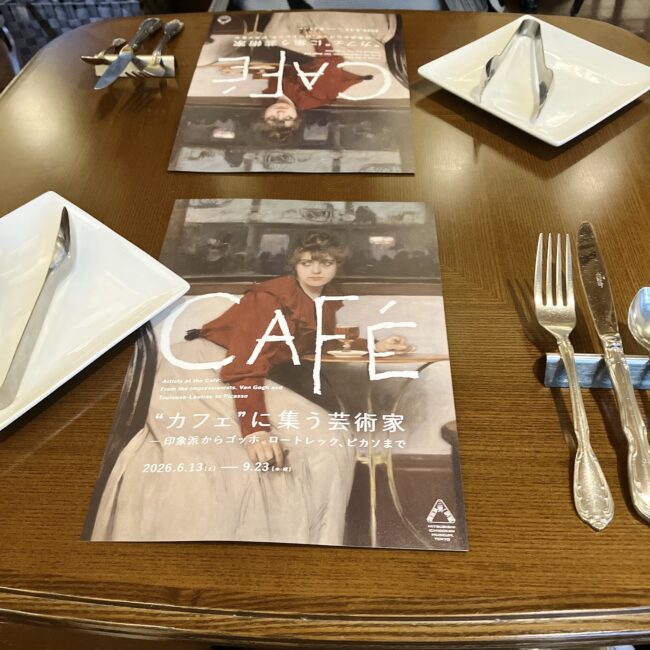 テーブルにセットされたカトラリーと、アフタヌーンティーのテーマである「“カフェ”に集う芸術家」展のチラシ。