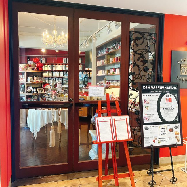 ガラス扉越しにシャンデリアや赤い紅茶缶が並ぶ可愛らしい店内が見える、デンメアティーハウス六本木店の入り口とメニュー看板。