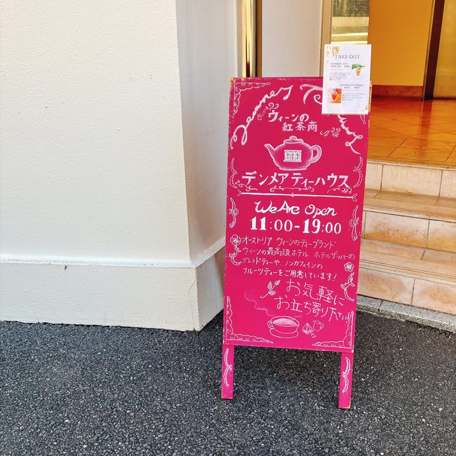 「ウィーンの紅茶商 デンメアティーハウス」と書かれた目を引くピンク色の立て看板。ティーポットの可愛いイラストや営業時間が描かれている。